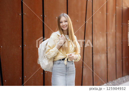 Young woman with long hair and backpack smiles in front of modern urban architecture during daytime 125984932