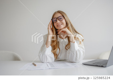 Young woman working at desk with laptop and documents while smiling in bright, modern office setting Young woman working at desk with laptop and documents while smiling in bright, modern office setting 125985224
