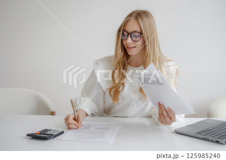 Young woman reviewing documents and writing notes in a bright home office setting with a laptop and calculator 125985240