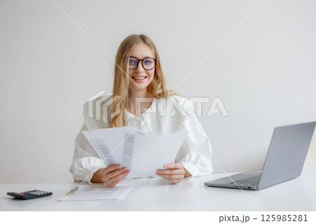 Woman reviewing documents at a desk with a laptop in a bright office environment during the day while smiling 125985281