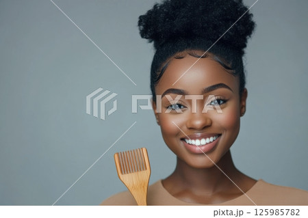 Young woman smiles brightly while holding a wooden hair tool against a plain background during a bright indoor setting 125985782