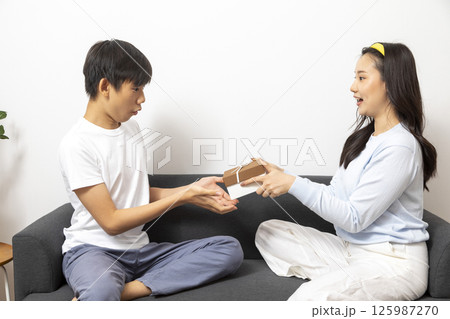 Excited, surprised woman receiving gift box from man sitting on a couch in the living room at home. Excited, surprised woman receiving gift box from man sitting on a couch in the living room at home. 125987270
