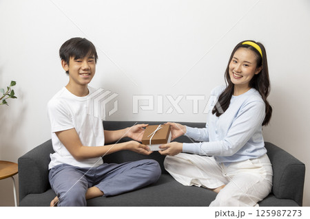 Excited, surprised woman receiving gift box from man sitting on a couch in the living room at home. 125987273