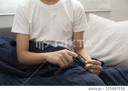 Sick asian teen boy with pulse oximeter on his finger. Ill child lying on sofa with blanket in living room. Sick asian teen boy with pulse oximeter on his finger. Ill child lying on sofa with blanket in living room. 125987558