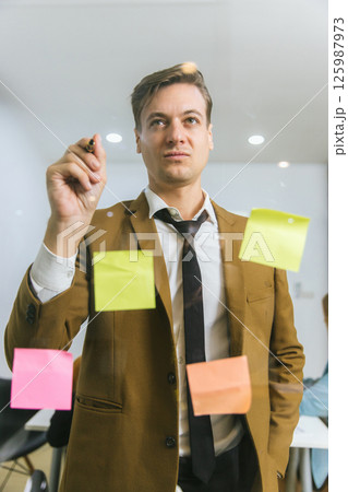 Business people holding pen and put sticky notes on glass wall in office Business people holding pen and put sticky notes on glass wall in office 125987973