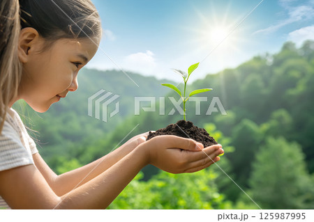 太陽をバックに森の前で緑の幼木を土と共に両手ですくいあげた女の子 125987995