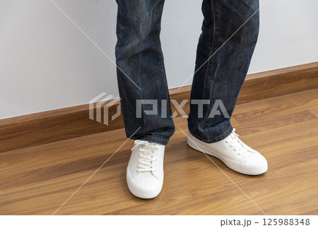 Feet wearing white chunky sole sneakers and blue jeans on the white wood floor Feet wearing white chunky sole sneakers and blue jeans on the white wood floor 125988348