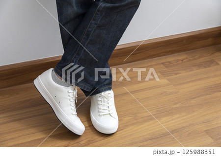 Feet wearing white chunky sole sneakers and blue jeans on the white wood floor Feet wearing white chunky sole sneakers and blue jeans on the white wood floor 125988351