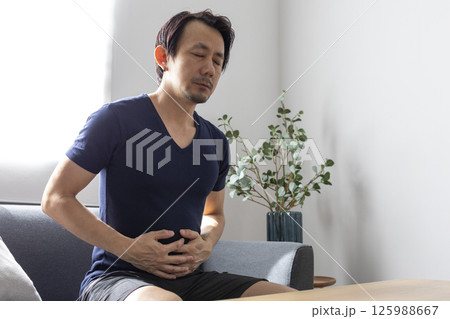 Mature 50s Asian man stomachache, pressing on stomach with painful expression, sitting on sofa at home, medicines on table. 125988667