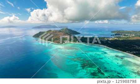 A lush green island stretches along the coast, with colorful coral reefs visible beneath turquoise waters. Praslin, Seychelles. 125988798