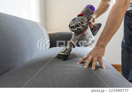 Adult Man in jeans and T-shirt vacuuming a sofa 125990777