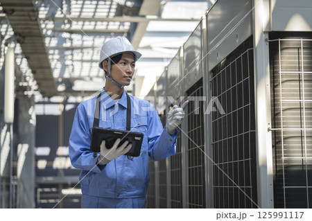 ビル屋上に設置された空調の室外機を点検する作業服姿の男性　ビルメンテナンス　イメージ　 125991117