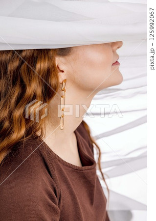 woman with long wavy red hair and golden earrings under white sheer fabric in brown open-back dress, feminine mood in studio scene, fashion, jewelry design, beauty editorial 125992067