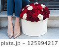 A luxurious bouquet of red and white roses is in a decorative gift box beside a stylishly woman legs in sparkly shoes. Valentines Day, flower store, shop, mock-up, delivery flowers, blog posts 125994217