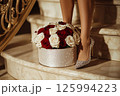 A luxurious bouquet of red and white roses is in a decorative gift box beside a stylishly woman legs in sparkly shoes. Valentines Day, flower store, shop, mock-up, delivery flowers, blog posts 125994223