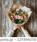 Florist hands hold bouquet of flowers wrapped in craft paper on wooden background. Flower delivery, flower store, shop advertising, blog posts, sale, shopping flowers. 125994250