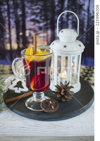 Mulled wine with orange and spices, Christmas decor and burning candles, on a white wooden table against the background of winter nature. Mulled wine with orange and spices, Christmas decor and burning candles, on a white wooden table against the background of winter nature. 125996208