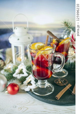 Mulled wine with orange and spices, Christmas decor and burning candles, on a white wooden table against the background of winter nature. Mulled wine with orange and spices, Christmas decor and burning candles, on a white wooden table against the background of winter nature. 125996210