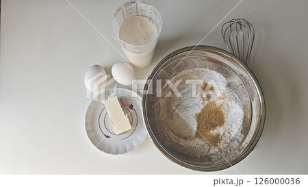 Ingredients for baking including flour, sugar, eggs, butter and milk arranged on a white table. Concept of homemade cooking, simple recipe preparation and traditional baking 126000036