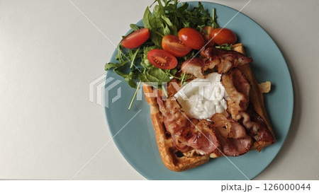Savory waffle breakfast with crispy bacon, poached egg, cherry tomatoes and arugula on blue plate. Concept of protein-rich brunch, healthy eating and modern food styling Savory waffle breakfast with crispy bacon, poached egg, cherry tomatoes and arugula on blue plate. Concept of protein-rich brunch, healthy eating and modern food styling 126000044