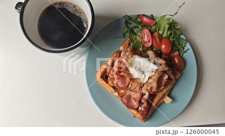 Waffle breakfast with crispy bacon, poached egg, arugula salad and a cup of black coffee. Concept of hearty morning meal, brunch comfort and savory food presentation Waffle breakfast with crispy bacon, poached egg, arugula salad and a cup of black coffee. Concept of hearty morning meal, brunch comfort and savory food presentation 126000045