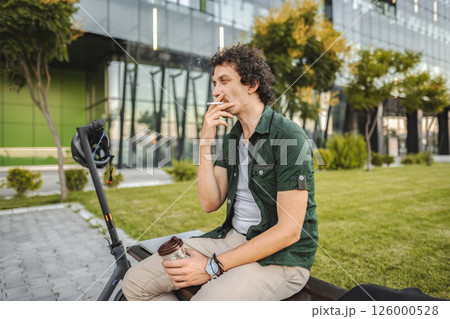 Man with curly hair sits on a park bench, smokes a cigar and rests 126000528
