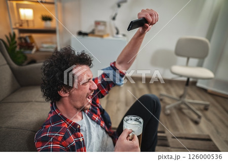 Excited sports fan cheering while watching tv and drinking beer Excited sports fan cheering while watching tv and drinking beer 126000536