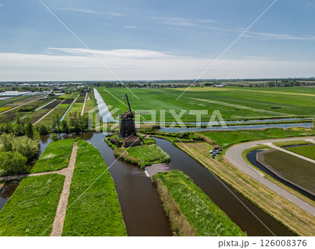 Aerial view of a Dutch countryside with a traditional windmill by a canal, green fields, and a nearby road with moving cars under a clear blue sky. Aerial view of a Dutch countryside with a traditional windmill by a canal, green fields, and a nearby road with moving cars under a clear blue sky. 126008376