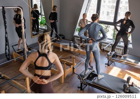 Group of caucasian women exercising with trainer on pilates reformers in a bright studio. Their workout focuses on core strength and flexibility in a modern fitness environment Group of caucasian women exercising with trainer on pilates reformers in a bright studio. Their workout focuses on core strength and flexibility in a modern fitness environment 126009511