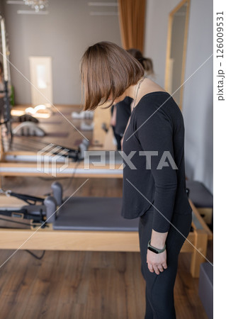 Young caucasian woman doing roll down exercise in a pilates bright studio. Their workout focuses on core strength and flexibility in a modern fitness environment. 126009531