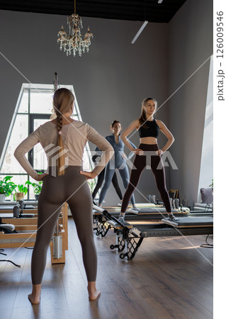 Young caucasian women exercising on pilates reformers in a bright studio. Their workout focuses on core strength and flexibility in a modern fitness environment. 126009546