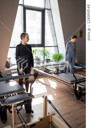 Young caucasian woman doing roll down exercise in a pilates bright studio. Their workout focuses on core strength and flexibility in a modern fitness environment. 126009548