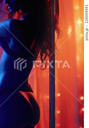 Silhouette of exotic dancer with long curly hair standing on pole in front of orange lights, backlit and cinematic. Silhouette of exotic dancer with long curly hair standing on pole in front of orange lights, backlit and cinematic. 126009941