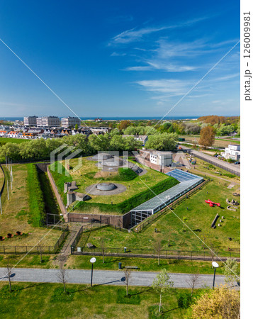 Aerial view of a historic military fort with grass-covered bunkers, surrounded by modern buildings, trees, and walking paths, near the coastline under a bright blue sky. 126009981