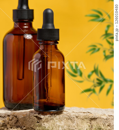 Amber glass dropper bottles on a natural stone surface with a yellow background and green plant elements. 126010480