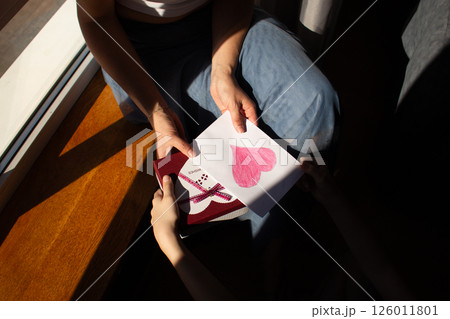 Young woman receiving gift box and handmade greeting card with painting from daughter for mother's day, 8 march, birthday. Close up. 126011801