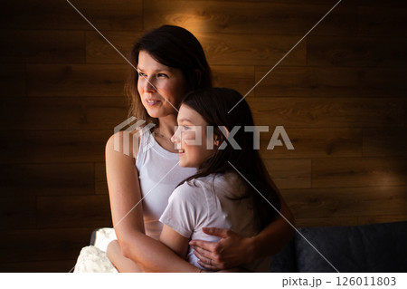 Adoptive mother holds her daughter close standing near window showing connection and warmth. Concept of modern family and power of love beyond biology. Adoptive mother holds her daughter close standing near window showing connection and warmth. Concept of modern family and power of love beyond biology. 126011803