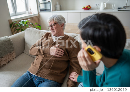 Elderly man clutching chest while sitting on chair, wife calls emergency services with phone nearby  126012059