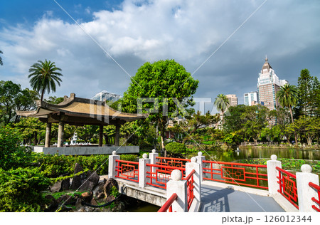 Traditional Chinese-style pavilion and red ornamental bridge sit peacefully in Zhongshan Park park in Taipei, Taiwan, with a reflective pond and modern buildings in the background under dramatic skies 126012344