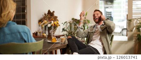 Lifestyle portrait of handsome young man, sitting in cafe, talking to a friend on mobile phone with wireless earphones, laughing and smiling, listening to funny podcast 126013741