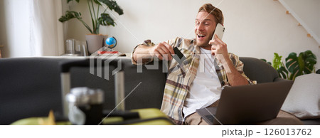 Image of guy calling someone, holding credit card and using laptop, has suitcase and camera packed for holiday 126013762