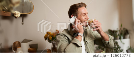 Portrait of young happy man, answers phone call, talking on mobile, drinks coffee in cafe, enjoys his free time Portrait of young happy man, answers phone call, talking on mobile, drinks coffee in cafe, enjoys his free time 126013778