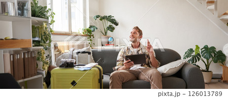 Excited young with tablet, sits in living room with packed suitcase, cross fingers for good luck, making wish, hoping for something Excited young with tablet, sits in living room with packed suitcase, cross fingers for good luck, making wish, hoping for something 126013789