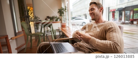 Portrait of handsome young man sits in cafe, drinks coffee and watches video on laptop, looking at screen with happy smile, relaxing in co-working space Portrait of handsome young man sits in cafe, drinks coffee and watches video on laptop, looking at screen with happy smile, relaxing in co-working space 126013885