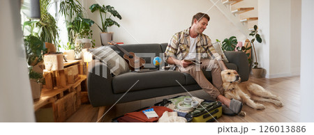 Man with a dog sitting near suitcase, holding notebook, checking list of items to take on vacation, planning a holiday trip 126013886