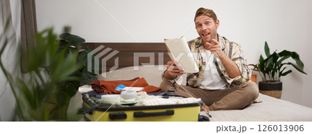 Portrait of happy, charismatic young man, excited about holiday, holding notebook, packing suitcase with clothes for vacation, pointing finger at camera and smiling 126013906