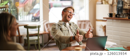 Portrait of satisfied, happy young man in glasses, working in cafe, sitting in co-working space with laptop, showing thumbs up, like and approve smth good, chatting, giving online lessons Portrait of satisfied, happy young man in glasses, working in cafe, sitting in co-working space with laptop, showing thumbs up, like and approve smth good, chatting, giving online lessons 126013934