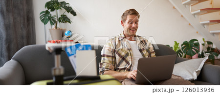 Portrait of happy young man booking holiday for two, has plane tickets, looks at laptop, sits in living room with computer Portrait of happy young man booking holiday for two, has plane tickets, looks at laptop, sits in living room with computer 126013936