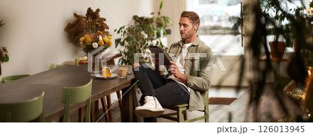 Lifestyle shot of handsome young man sitting in a cafe in front of the table, drinking coffee, wearing wireless headphones, listening to music or watching video in public space, using digital tablet 126013945