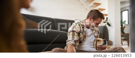 Happy young smiling guy in living room, sitting on floor, hugging his pet, playing with dog 126013953